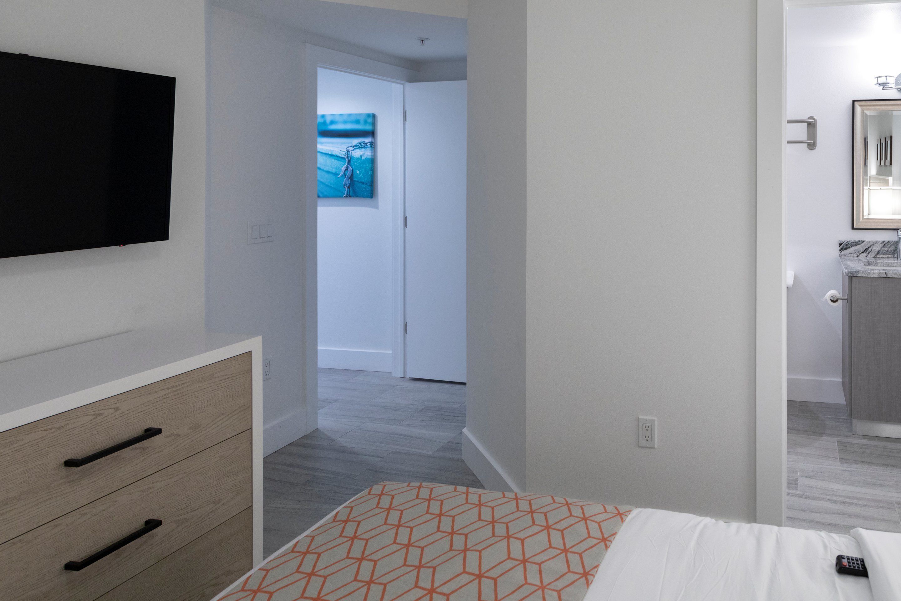 A hotel room with a bed, a TV mounted on the wall, a wooden dresser, and a bathroom visible through an open door.