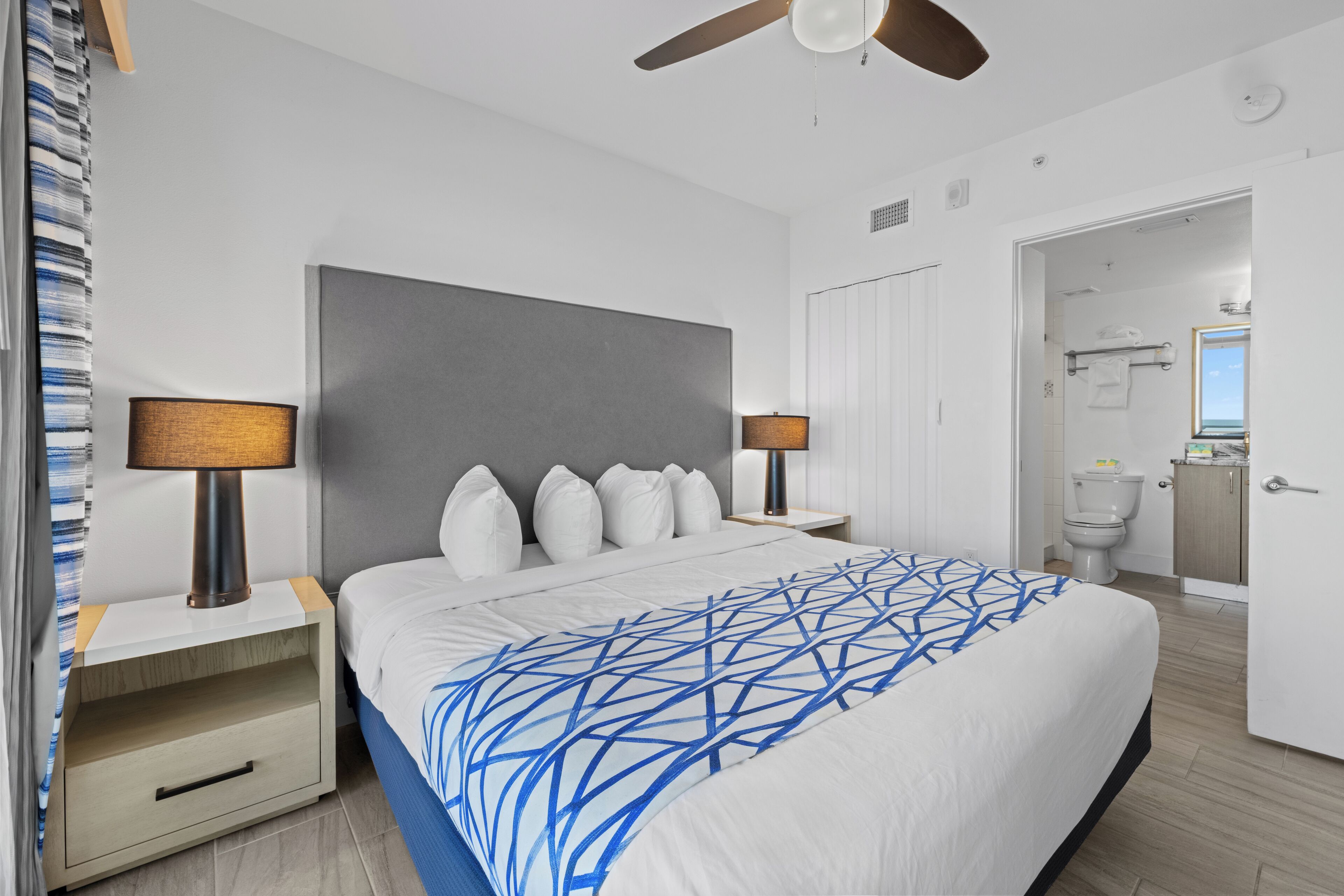 A modern hotel room with a large bed, two bedside lamps, a ceiling fan, and a bathroom visible through an open door.