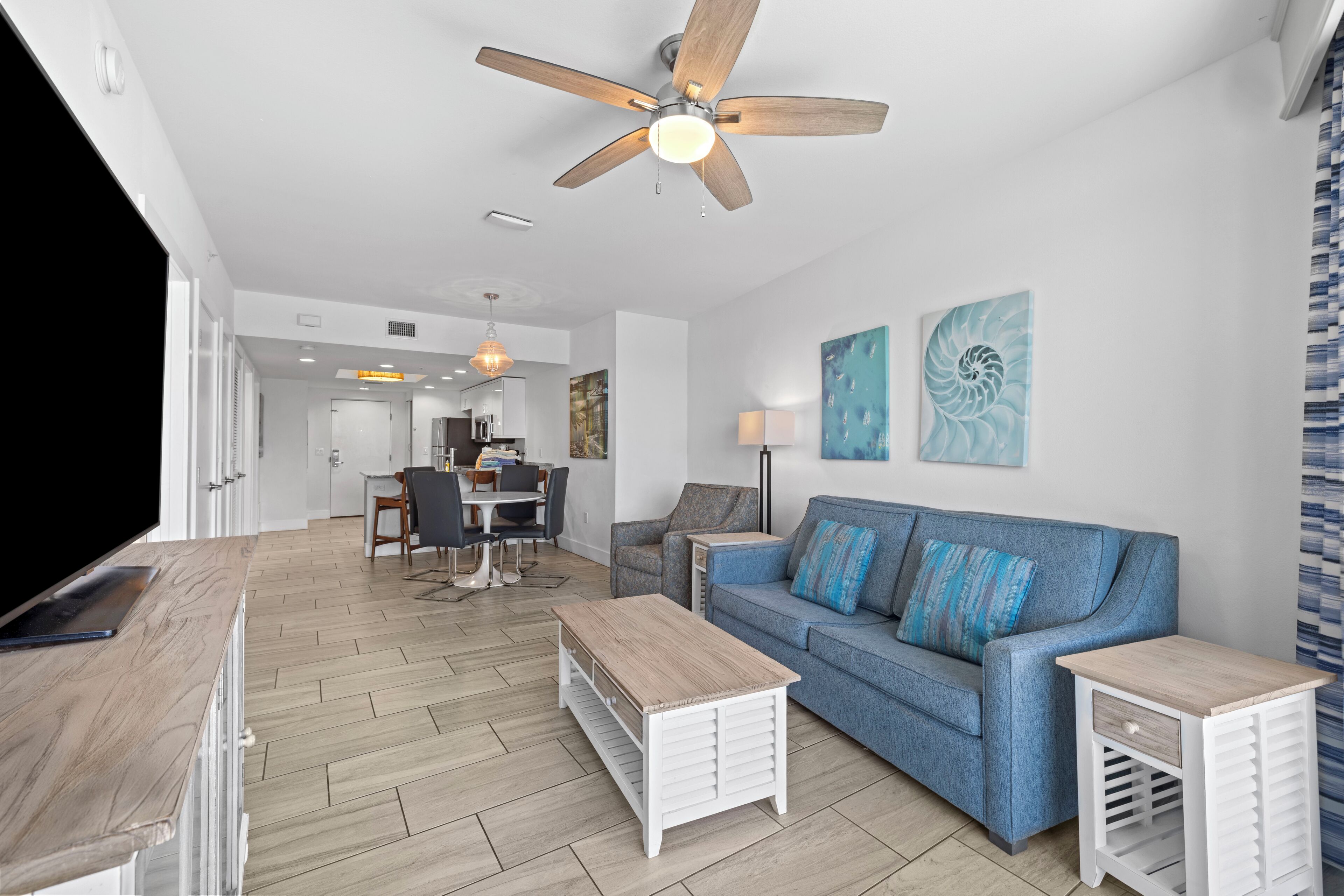 A modern living room with a blue sofa, white side tables, and a wooden coffee table. There is a ceiling fan, a TV, and artwork on the walls.