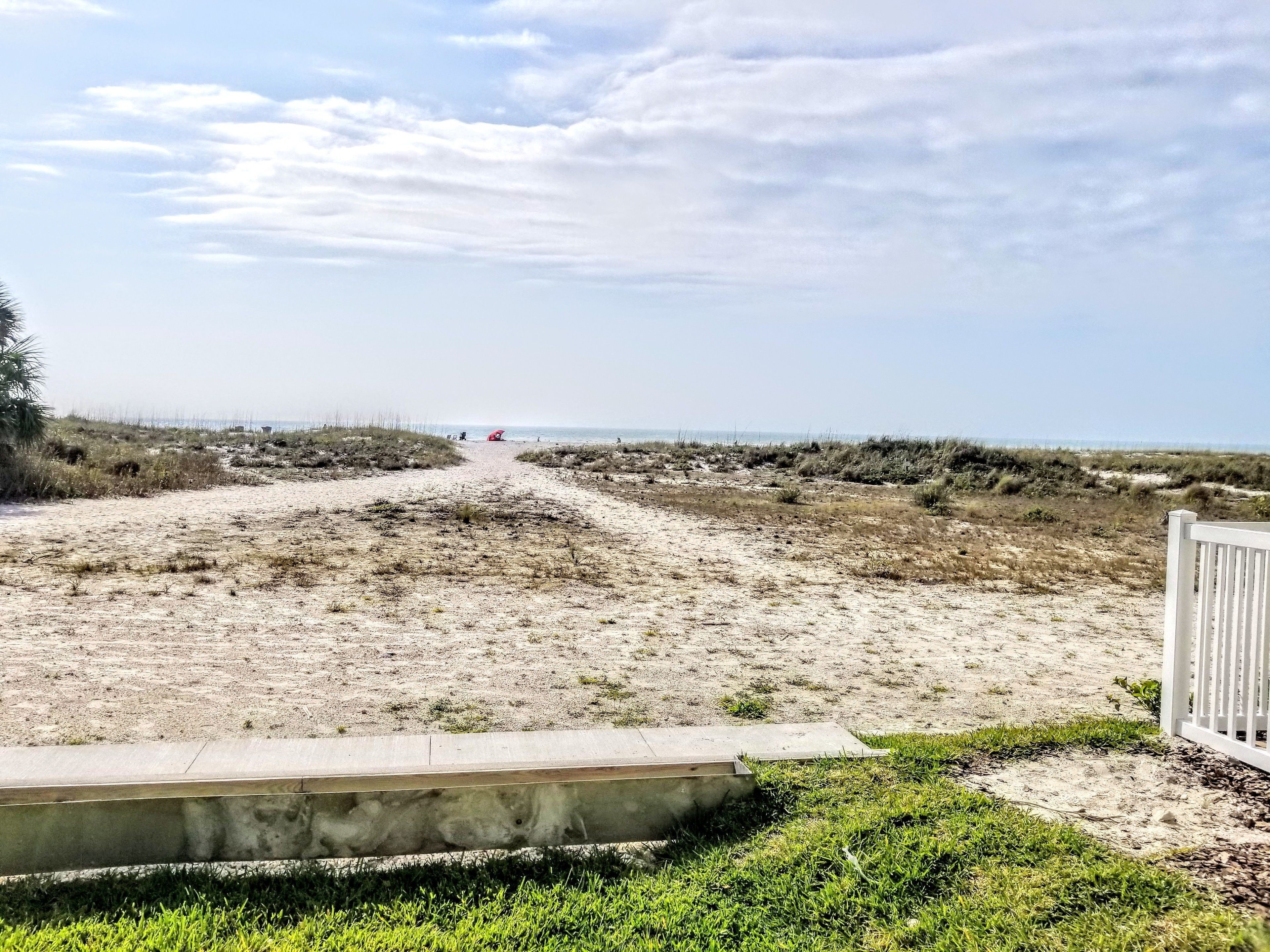 A sandy beach path leading to the sea with a white fence on the right.
