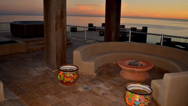 A rooftop terrace with a hot tub, two decorative pots, and a view of the ocean at sunset.