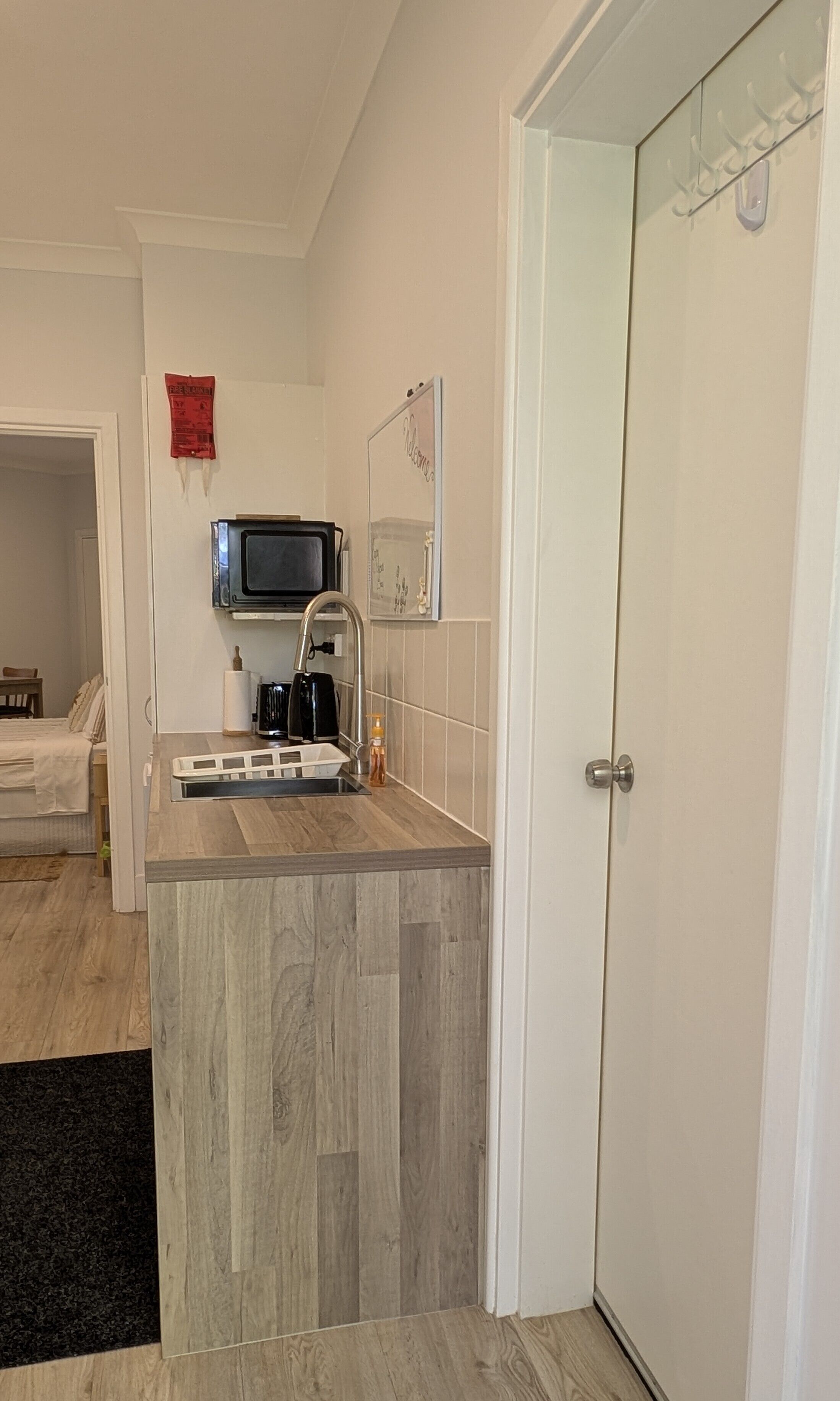 Een kleine keuken met een inbouwmagnetron, koffiezetapparaat en een spoelbak. Er is een deur met een kapstok en een welkomstbord. Een bed is zichtbaar in de aangrenzende kamer.