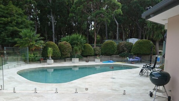 A swimming pool with a curved edge, surrounded by a tiled patio. There are potted plants and a barbecue grill. - Amberlyn Escapes Killcare (Killcare Heights)