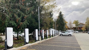 Un estacionamiento con múltiples estaciones de carga para Tesla, un coche blanco estacionado en el lugar, y un edificio con techo rojo al fondo.