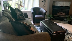 Una sala de estar con sofá, sillón y reposapiés, chimenea y un ventanal con persianas.