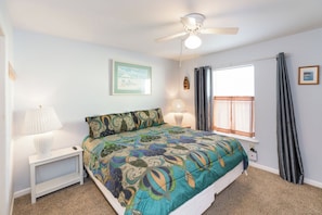 A bedroom with a bed, a nightstand, a lamp, a ceiling fan, a window with curtains, and a framed picture on the wall. - Shell Shocked - Oceanfront Surf Condo, Sleeps 6 (Surf City)