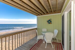 A wooden deck with a view of the beach and ocean. There is a table with chairs and a decorative wall hanging. - Shell Shocked - Oceanfront Surf Condo, Sleeps 6 (Surf City)
