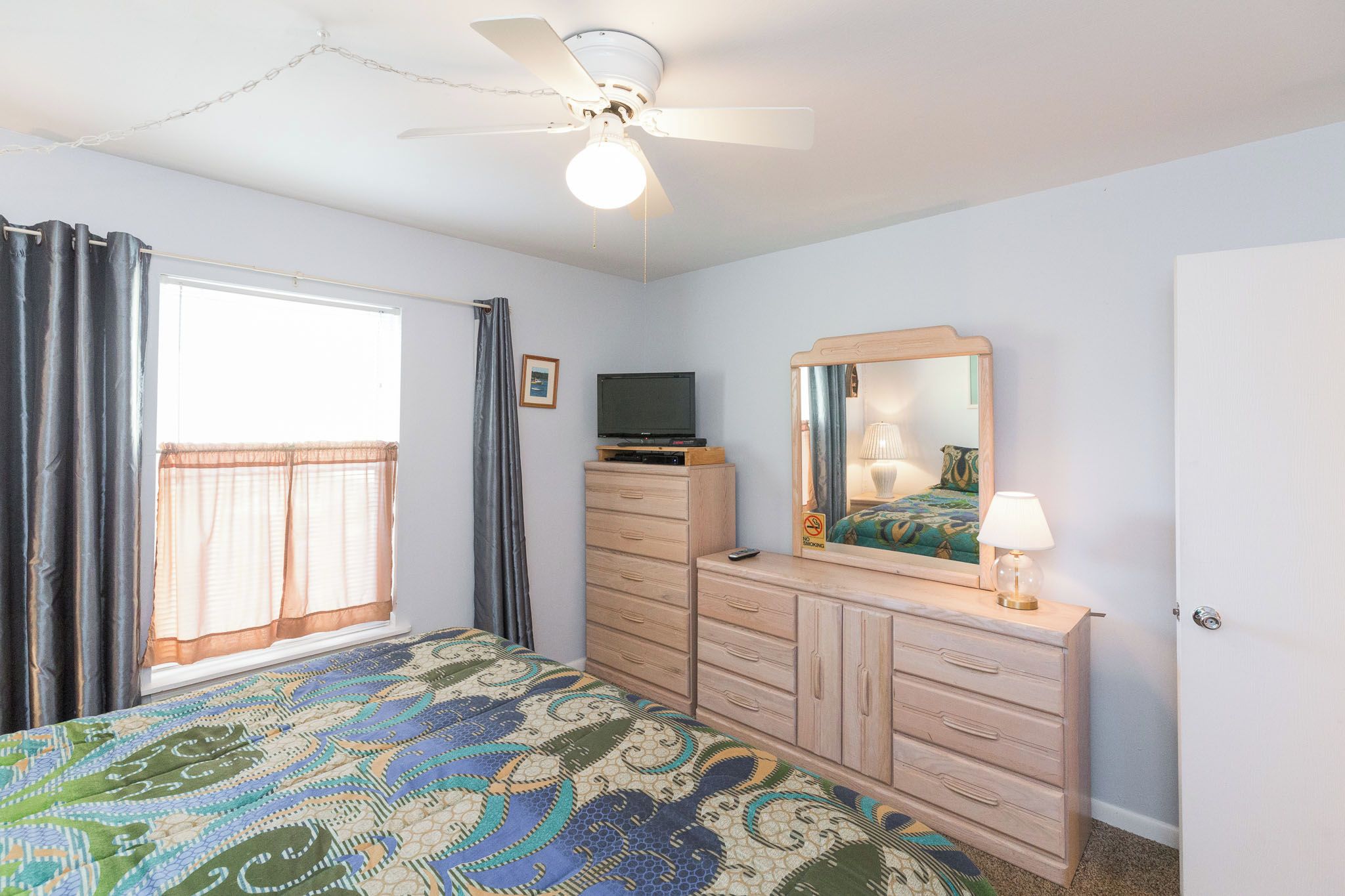 Une chambre à coucher comprenant un lit, une commode, un téléviseur, un ventilateur de plafond et une fenêtre avec des rideaux.