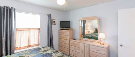 A bedroom with a bed, a dresser, a television, a ceiling fan, and a window with curtains.