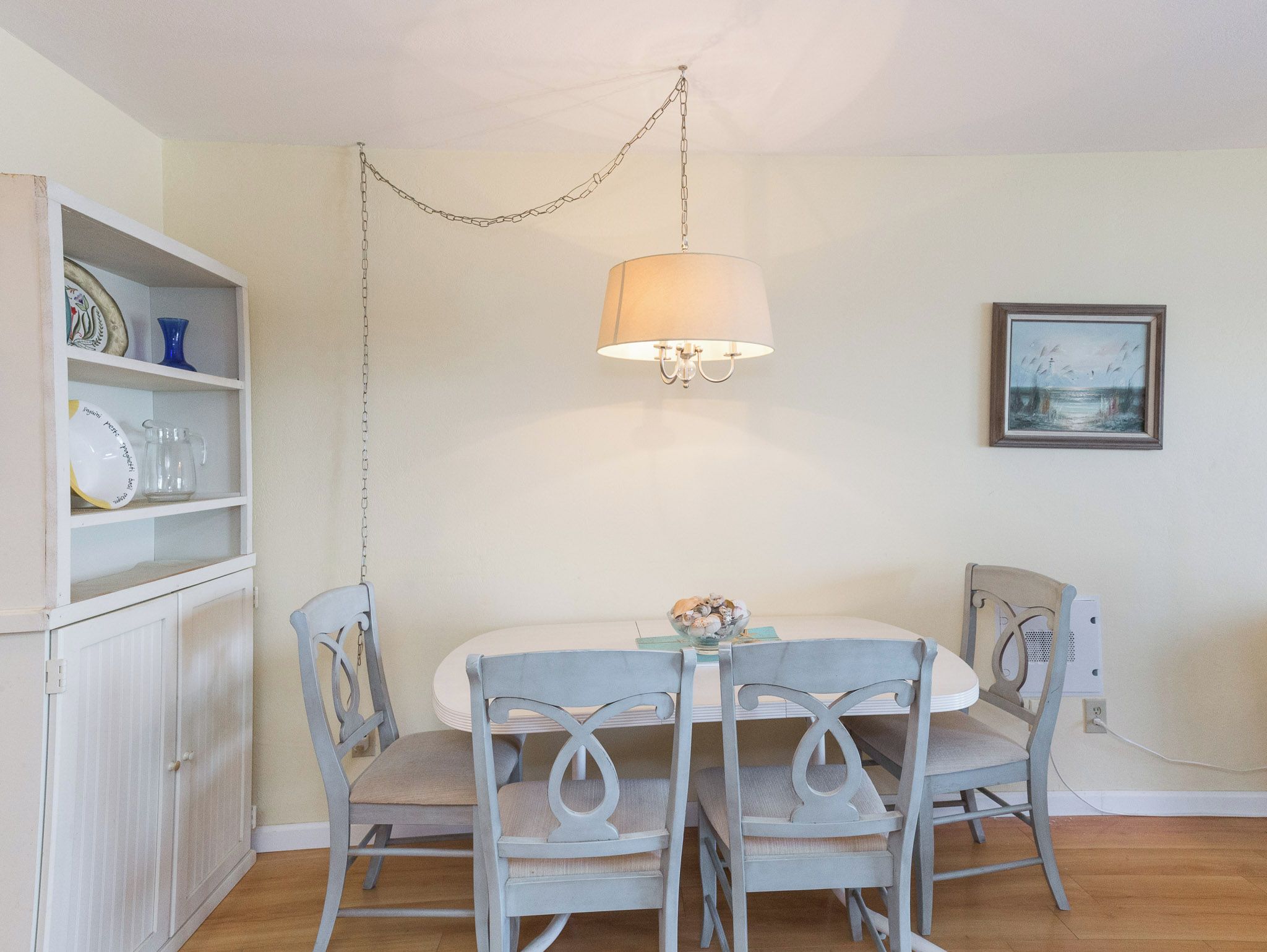 A dining area with a white table, four grey chairs, a hanging light fixture, and a wall-mounted picture.