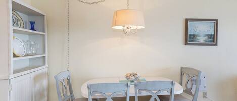 A dining area with a white table, four grey chairs, a hanging light fixture, and a wall-mounted picture.