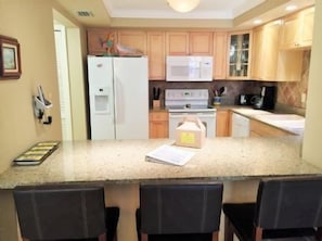 A kitchen with a central island, a refrigerator, a microwave, and a stove. There are chairs around the island.