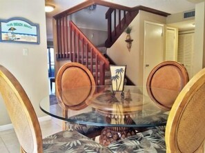 A dining area with a glass table surrounded by wicker chairs, a wooden staircase in the background, and a framed picture on the wall.