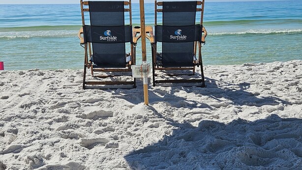 Zwei Strandstühle mit 'Surfside Resort'-Branding, ein großer blauer Sonnenschirm und ein Sandstrand mit dem Meer im Hintergrund.