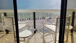 Ein Balkon mit weißen Stühlen und einem kleinen Tisch, mit Blick auf einen Strand mit Sonnenschirmen und Menschen.