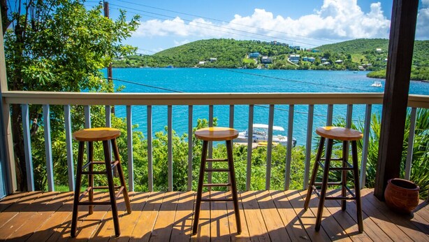 Une terrasse en bois avec trois tabourets, d’où l’on peut admirer une mer turquoise et des collines verdoyantes.