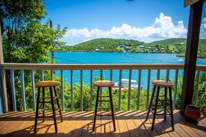 Eine Holzterrasse mit drei Hockstühlen mit Blick auf ein türkisblaues Meer und grüne Hügel.