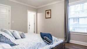 Un dormitorio con cama, un banco de madera, una puerta, una ventana con cortinas y un cuadro enmarcado en la pared.