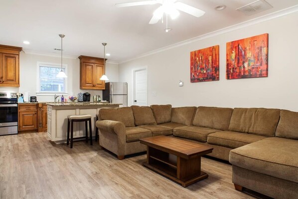 A modern living room with a sectional sofa, a wooden coffee table, and a kitchen area with wooden cabinets and a refrigerator. - Heart of Midtown - Walkable - Private Pet Relief Yard - Gated Parking (Atlanta)