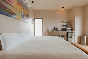 Habitació d'hotel amb un llit gran, un quadre a la paret, una cuineta i una porta que condueix a una altra habitació.