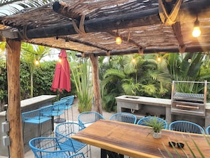 An outdoor patio with wooden tables and blue chairs, surrounded by lush greenery and a wooden pergola with hanging lights. - Vista Tortuga Ocean View VILLA-SUNSET Heated Pool by the BEACH  (Todos Santos)