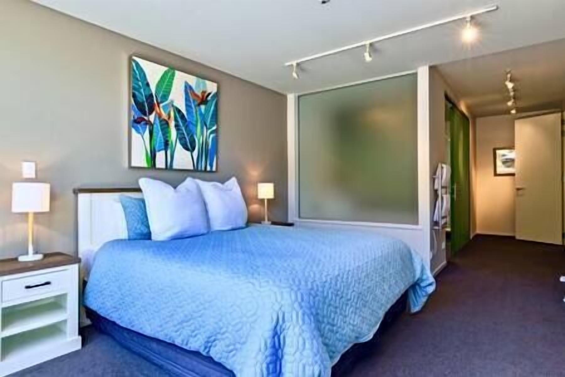 A modern hotel room with a large bed, a nightstand, a wall-mounted painting, and a sliding door leading to another room.