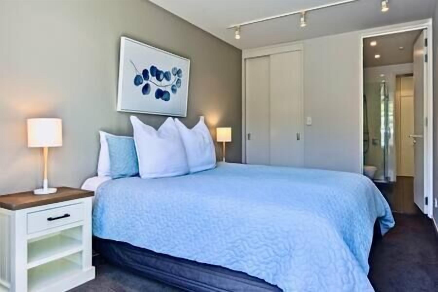 A bedroom with a bed, bedside lamps, a nightstand, a wall-mounted artwork, and a door leading to another room.