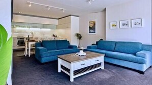 Ein modernes Wohnzimmer mit einem blauen Sofagarnitur, einem Holztisch und einer Küche im Hintergrund.