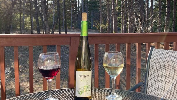 Une bouteille de vin Crestwood Barrel et deux verres sur une table en métal, à l’extérieur, avec une rambarde en bois et des arbres en arrière-plan.