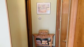 Un couloir étroit au plancher de bois, une petite table en bois sur laquelle sont posés des livres, et un tableau encadré accroché au mur.