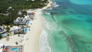 Vista aérea de un complejo turístico en la playa con varias piscinas, sombrillas y villas frente al mar.