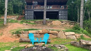 A cabin with a stone foundation, a wooden deck, and a fire pit with two chairs. - Appalachian Container Cabin (Otto)