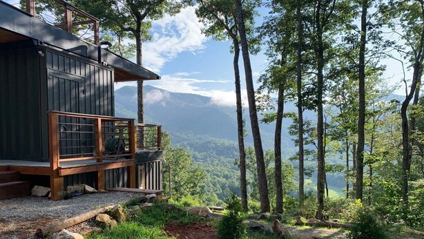 A cabin with a wooden deck and a glass enclosure overlooking a forested valley. - Appalachian Container Cabin (Otto)