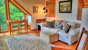Ein gemütliches Wohnzimmer mit einer Couch, Hocker und Holzmöbeln, ein Essbereich mit Stühlen und einem Tisch, sowie ein großes Fenster mit Blick auf Grünflächen.