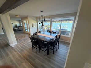 A dining area with a large table and chairs, a view of the water, and a living room in the background. - PNW & PLAY @ Lake Samish (Bellingham)