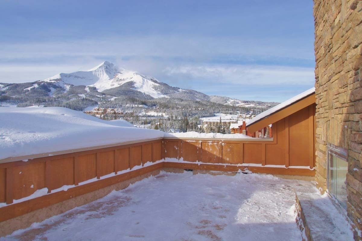 A snow-covered balcony with a mountain view, wooden railing, and stone wall.