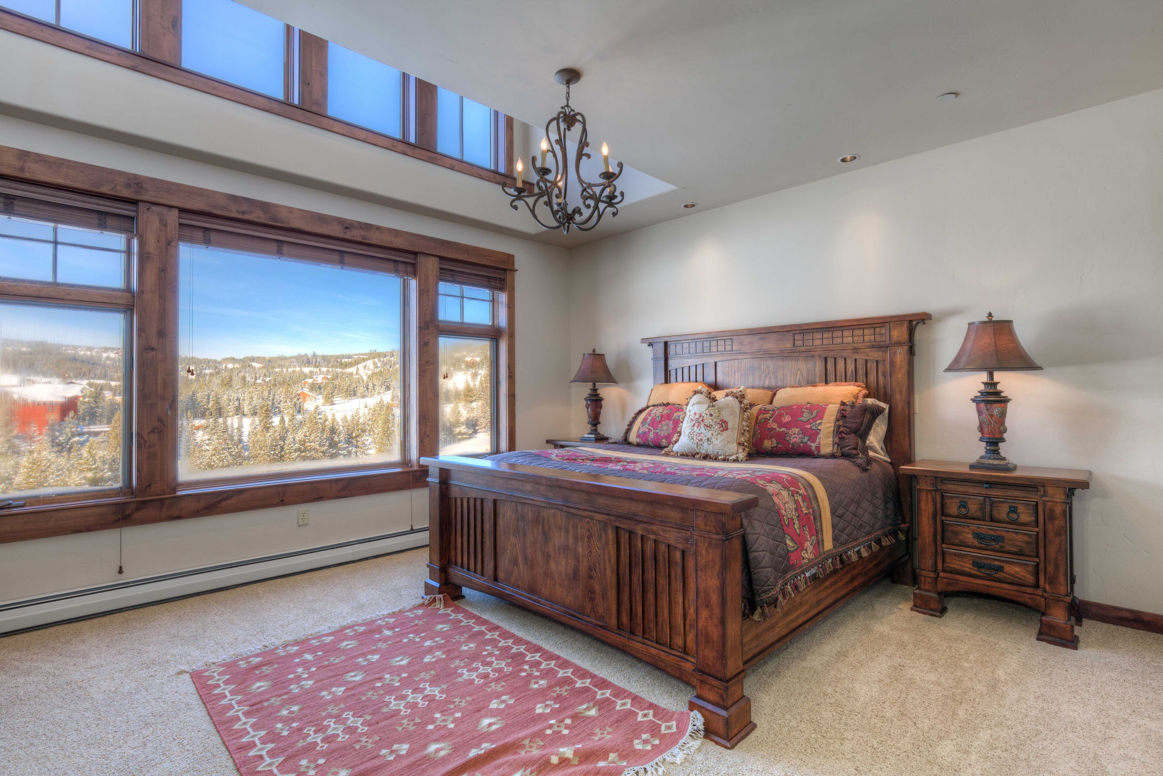 A bedroom with a large window, a bed with multiple pillows, a nightstand, and a chandelier.