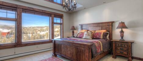 A bedroom with a large window, a bed with multiple pillows, a nightstand, and a chandelier.