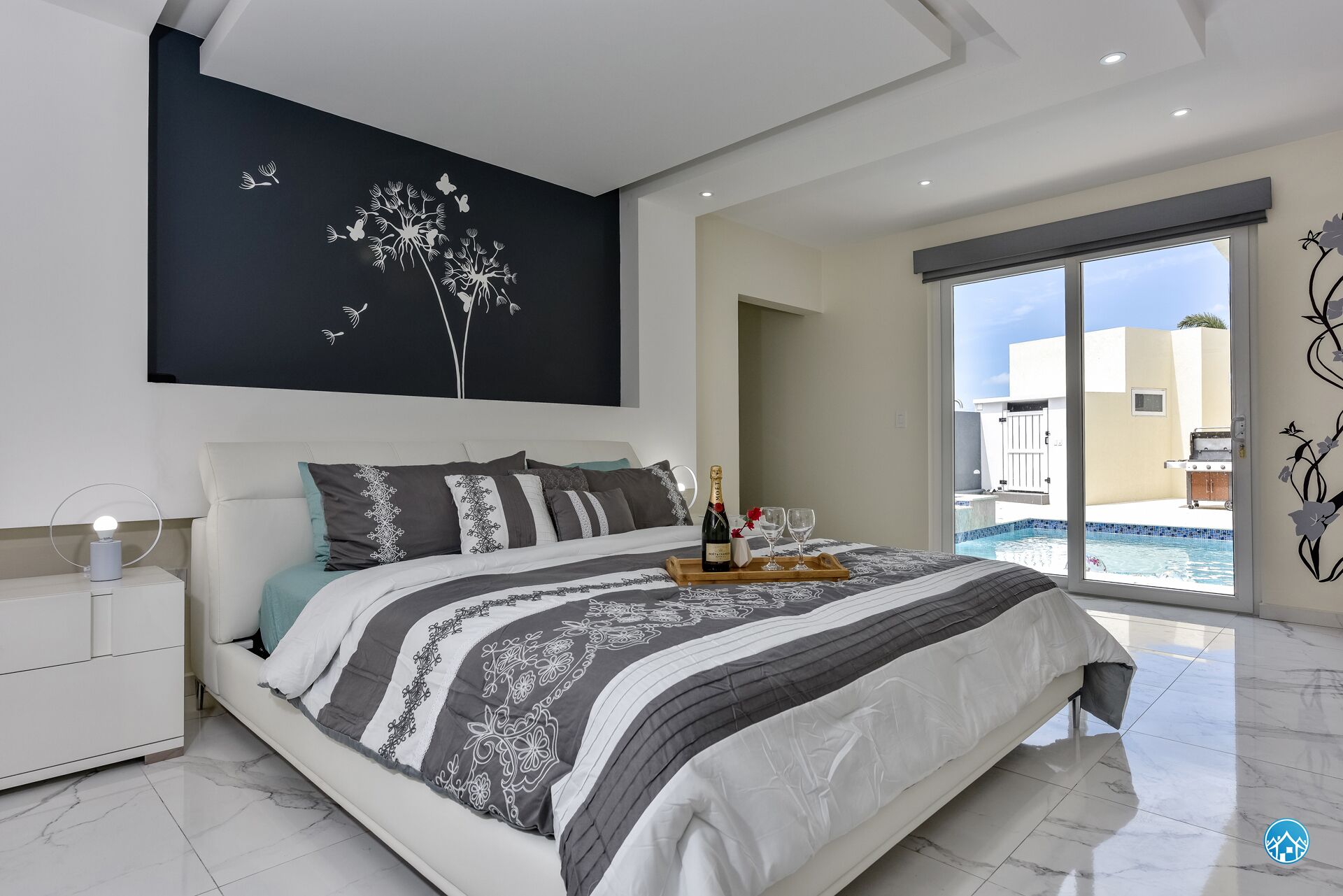 Une chambre moderne avec un grand lit, une table de chevet avec une lampe, un plateau avec une bouteille et des verres, et une vue sur une piscine par une porte coulissante.