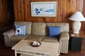 Um quarto com sofá bege, duas almofadas, mesa de centro de vime com cesta, um quadro na parede e um abajur sobre uma mesinha de madeira.