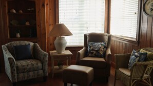 A living room with a patterned armchair, a cushioned ottoman, a side table with a lamp, and a bookshelf. - Oceanfront 4BR 2 Story Duplex with Private Beach Access (Carolina Beach)