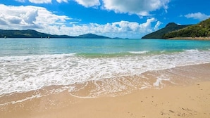 A beach with clear turquoise water, gentle waves, and a distant mountain range under a partly cloudy sky. - Frangipani Lodge 106, Beachfront Apartment on Hamilton Island (Hamilton Island)