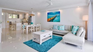 A modern living room with a white sofa, a coffee table, and a dining area with white chairs. - Frangipani Lodge 106, Beachfront Apartment on Hamilton Island (Hamilton Island)