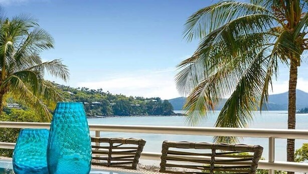 A balcony with wicker furniture, a glass table, and a view of a lake with palm trees and a distant mountain. - Frangipani Lodge 106, Beachfront Apartment on Hamilton Island (Hamilton Island)