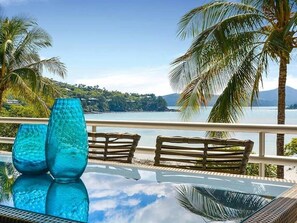 A balcony with wicker furniture, a glass table, and a view of a lake with palm trees and a distant mountain. - Frangipani Lodge 106, Beachfront Apartment on Hamilton Island (Hamilton Island)