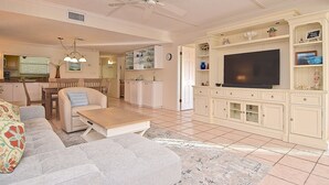 A living room with a sofa, coffee table, TV, and kitchen area with cabinets and a dining table. - Steps to Siesta Key Beach! Direct Access to Private Beach, Heated Pool. Sea Shell (Siesta Key)
