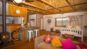 Ein gemütliches Wohnzimmer mit Kamin, Holzbalkendecke, einer Couch mit bunten Kissen, einer Küchenzeile mit Barhocker und einer Wand mit Traumfängern.