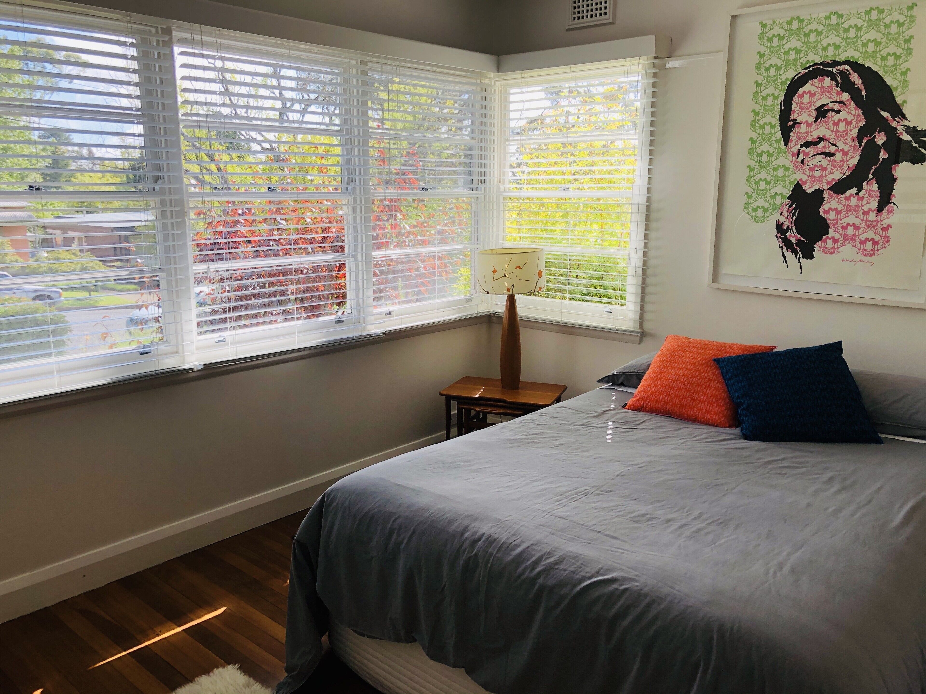 A bedroom with a bed, pillows, a nightstand, a lamp, and a window with blinds.