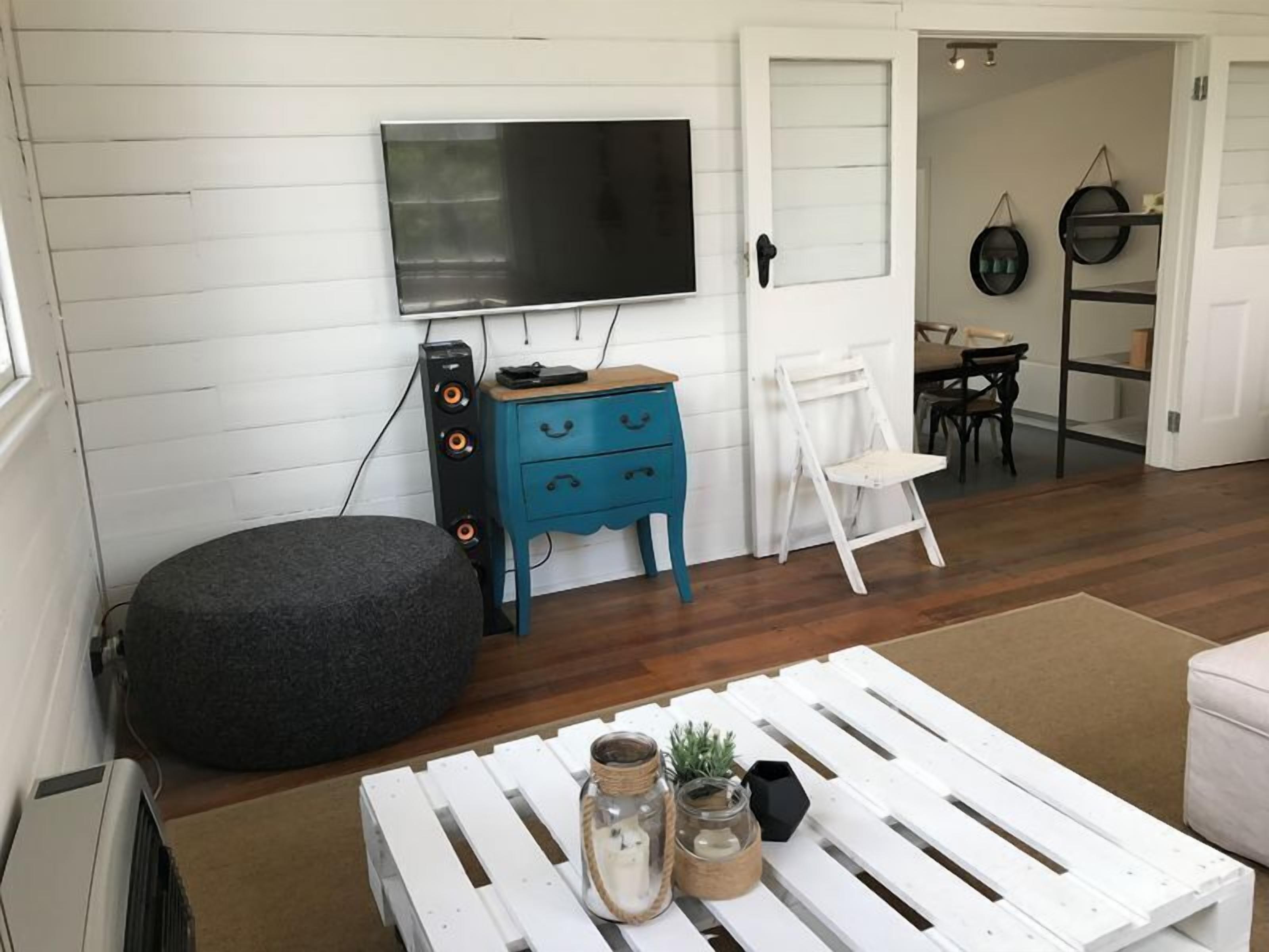 A living room with a flat-screen TV, a blue dresser, a white chair, a round ottoman, and a white coffee table with decorative jars.