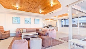 Een moderne woonkamer met een houten plafond, een hoekbank, poefjes, en een keuken op de achtergrond.